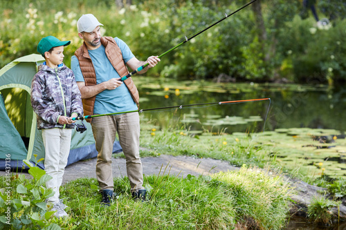 Full length portrait of loving father teaching son fishing while enjoying camping trip together, copy space