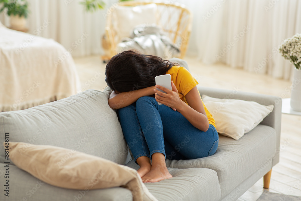 Obraz premium Sad young black woman with smartphone crying on sofa at home