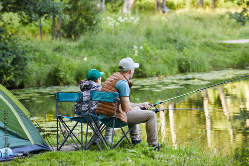 Side view full length portrait of loving father and son fishing by lake together during camping trip in nature, copy space