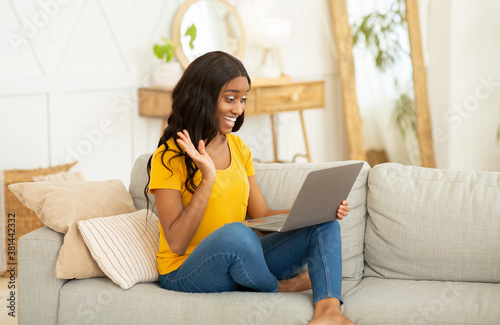 Online communication. Young black woman sitting on sofa at home and talking to family, friends or coleagues on laptop photo