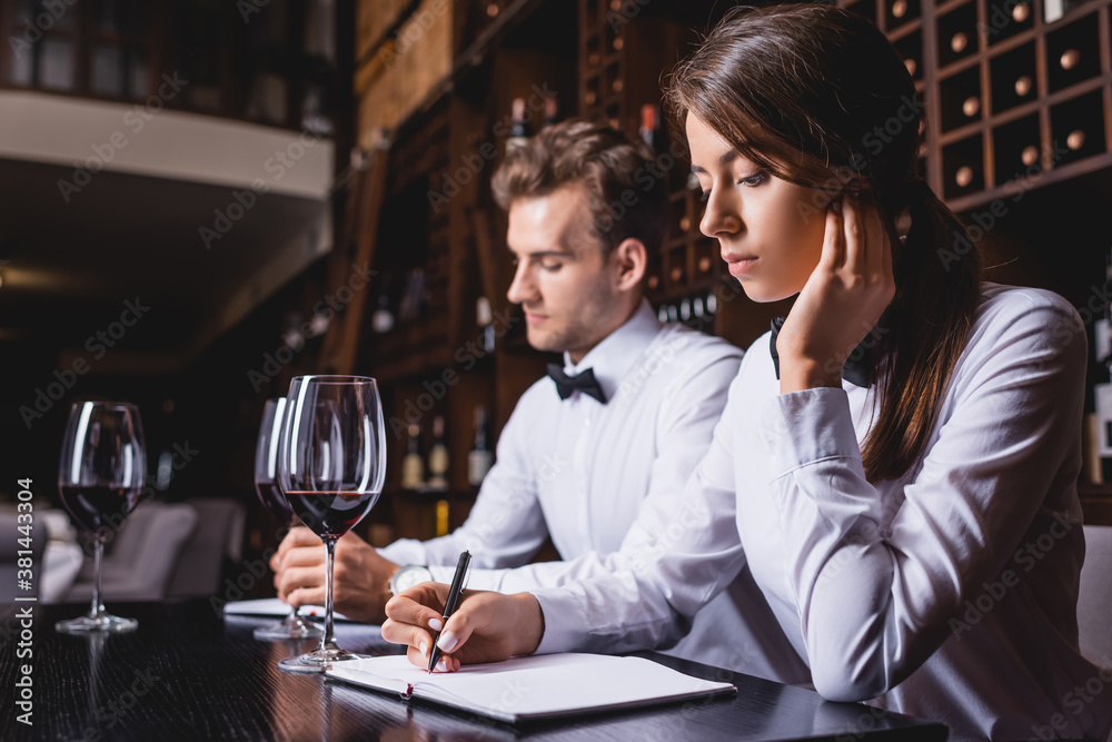 Selective focus of sommeliers writing on notebooks near glasses of wine in restaurant
