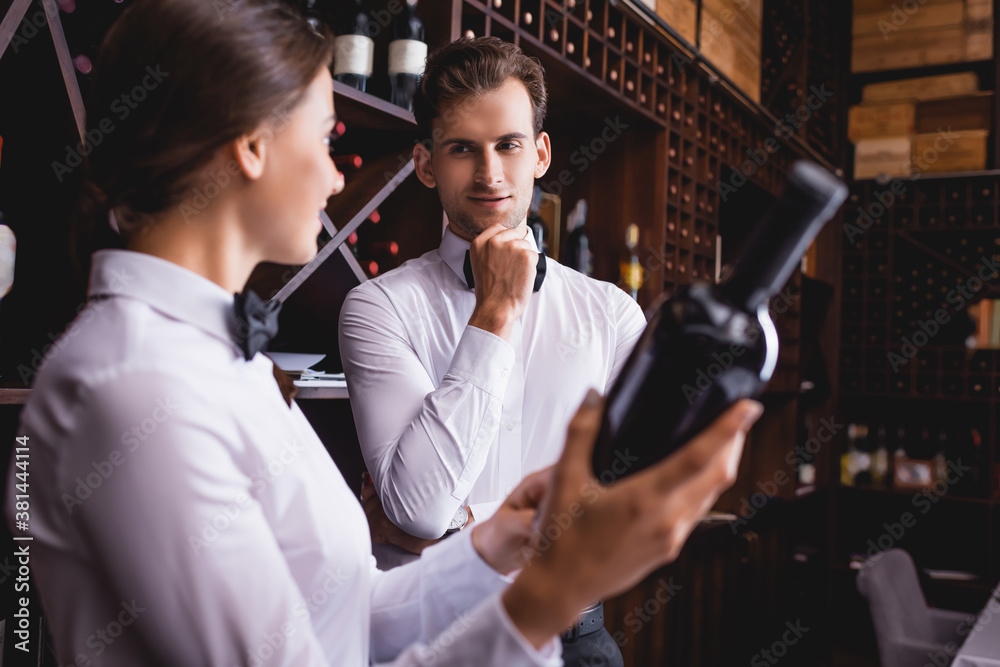 Selective focus of sommelier looking at colleague with bottle of wine in restaurant
