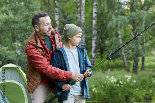 Side view portrait of mature father teaching son fishing while enjoying camping trip together by lake, copy space