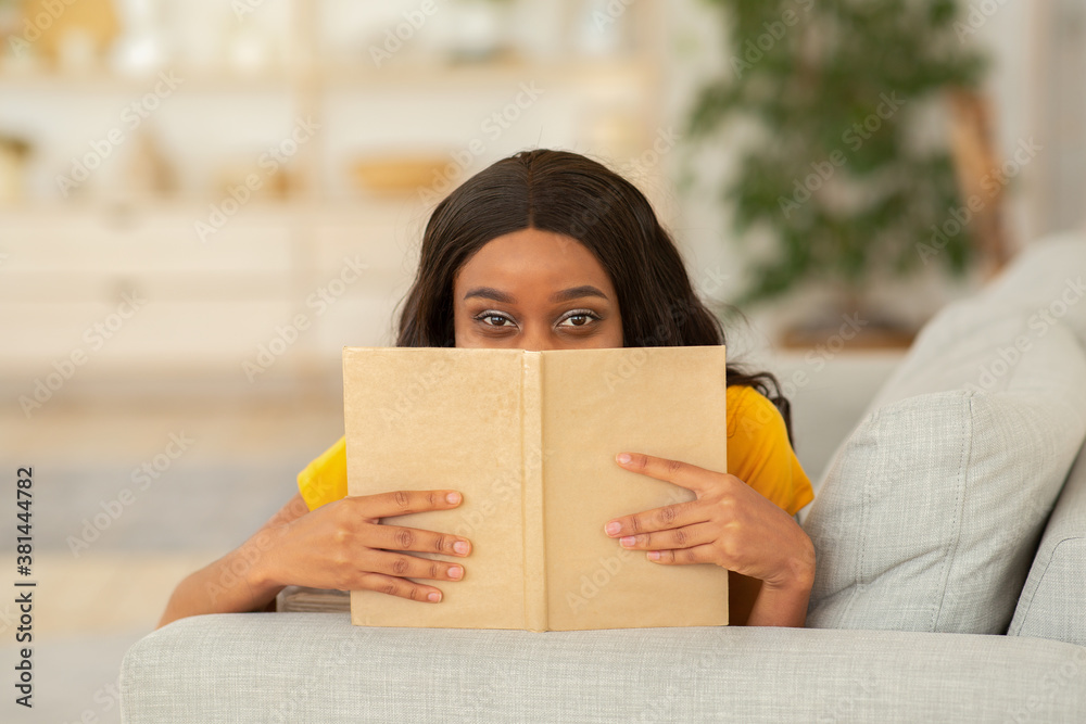 Stay home hobbies concept. Beautiful African American woman hiding behind book on sofa indoors Stay home hobbies concept. Beautiful African American woman hiding behind book on sofa indoors