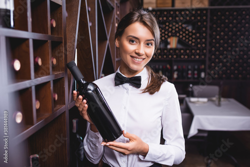 Selective focus of sommelier holding bottle of wine and looking at camera near racks photo