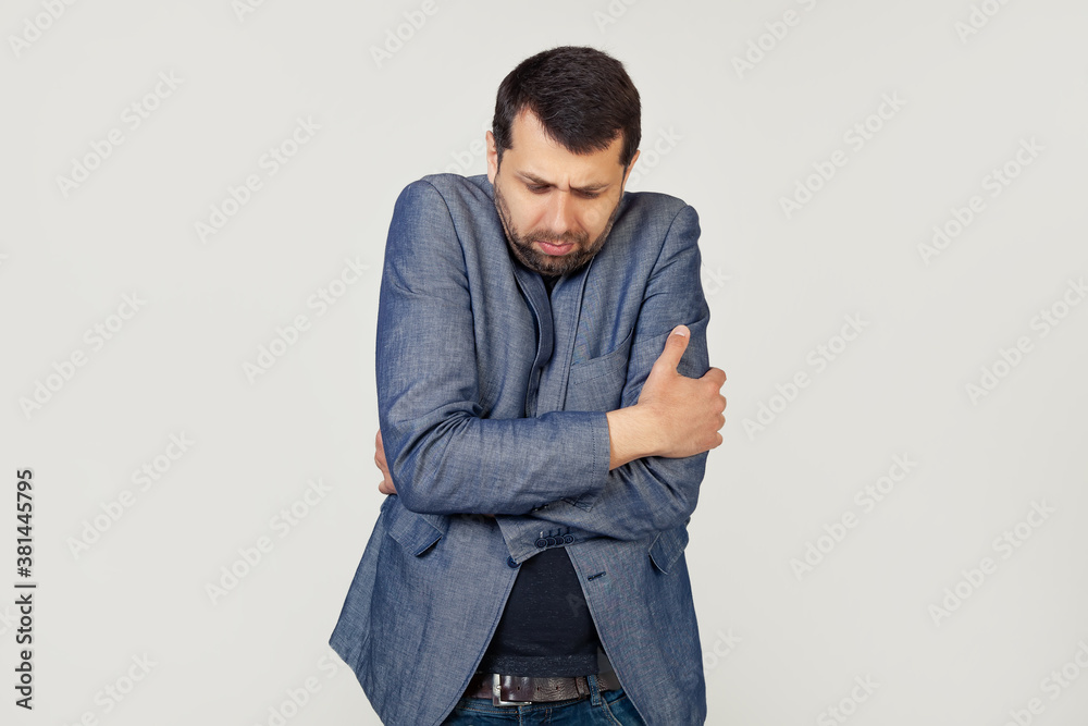 Young businessman man with a beard in a jacket, shaking and freezing from the winter cold with a sad and shocked expression on his face. Portrait of a man on a gray background