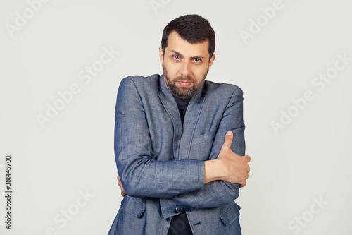 Young businessman man with a beard in a jacket, shaking and freezing from the winter cold with a sad and shocked expression on his face. Portrait of a man on a gray background © Yauhen