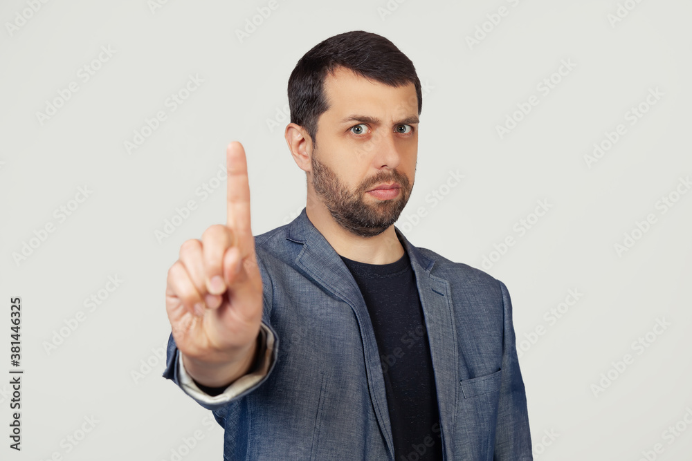 Young businessman man with a beard in a jacket pointing his finger up and angry expression without showing gestures. Portrait of a man on a gray background Young businessman man with a beard in a jacket pointing his finger up and angry expression without showing gestures. Portrait of a man on a gray background