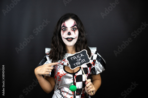 woman in a halloween clown costume over isolated black background holding blackboard with trick or treat word message