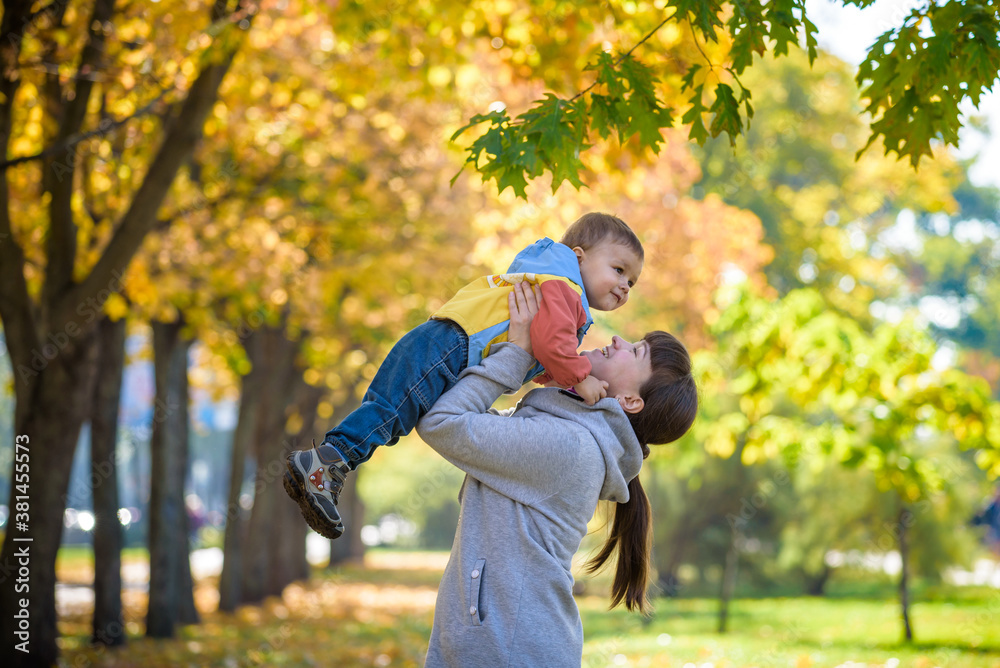 Fototapeta premium Happy young mother holding sweet toddler boy, family having fun together outside on a nice sunny autumn day. Cute adorable kid and mother play among maple leaves in autumn park or forest