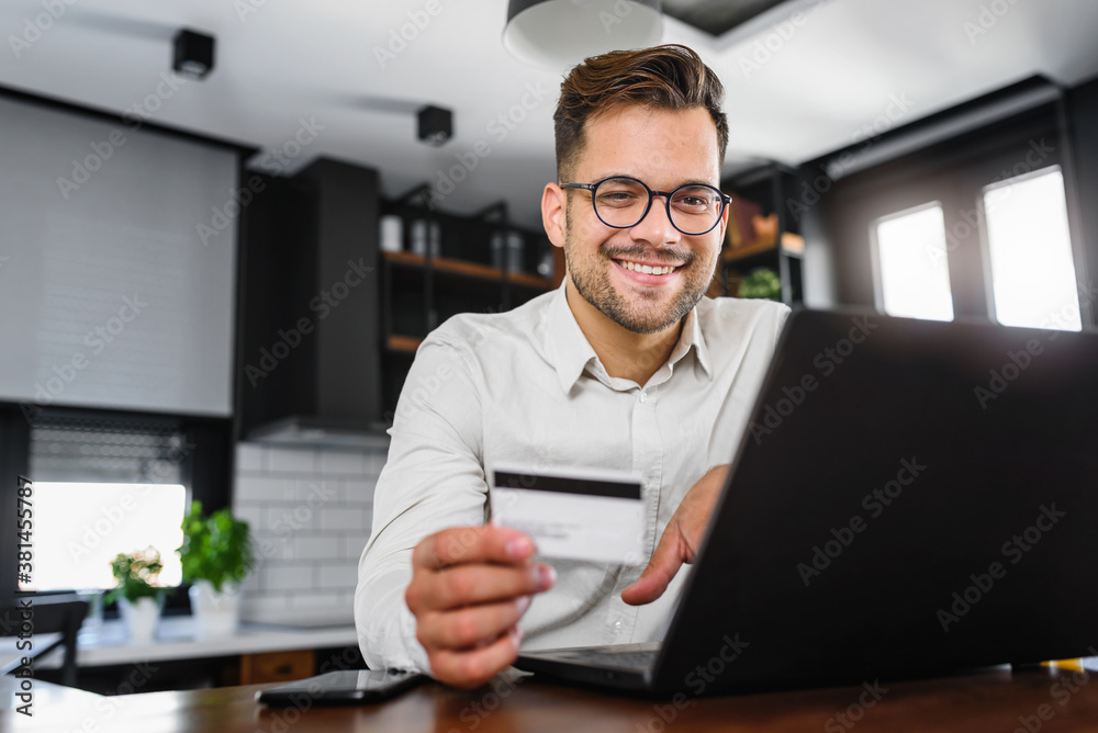Young man holding credit card sitting in front of laptop computer at home, paying for online order. People, lifestyle, modern technologies and e-commerce concept. Online banking and shopping using mod Young man holding credit card sitting in front of laptop computer at home, paying for online order. People, lifestyle, modern technologies and e-commerce concept. Online banking and shopping using mod