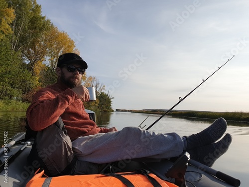 trolling fishing, a bearded man on a motor boat floating on the river on an autumn Sunny day and drinking tea from an iron mug.
