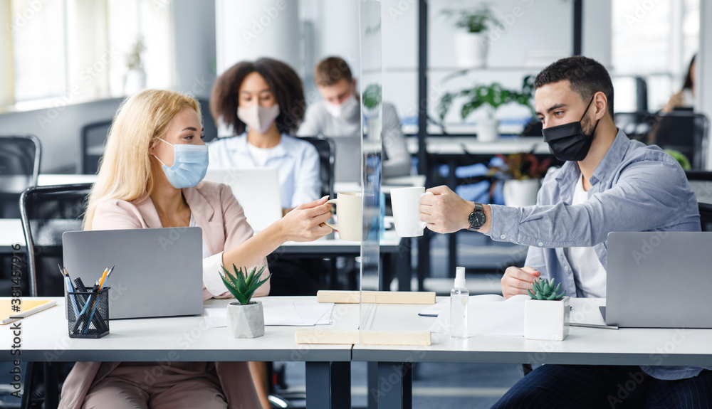 Coffee break in modern office and protecting health of workers. Young man and woman in protective masks clink cups through glass board in interior of office Coffee break in modern office and protecting health of workers. Young man and woman in protective masks clink cups through glass board in interior of office