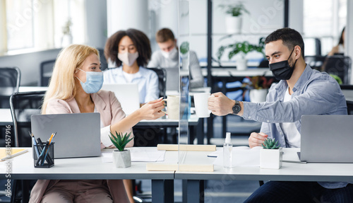 Coffee break in modern office and protecting health of workers. Young man and woman in protective masks clink cups through glass board in interior of office