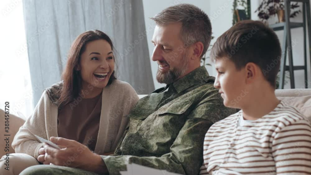 Handheld slowmo shot of cheerful male army veteran with beard sitting on couch and sharing deployment stories while showing photos to his happy wife and son