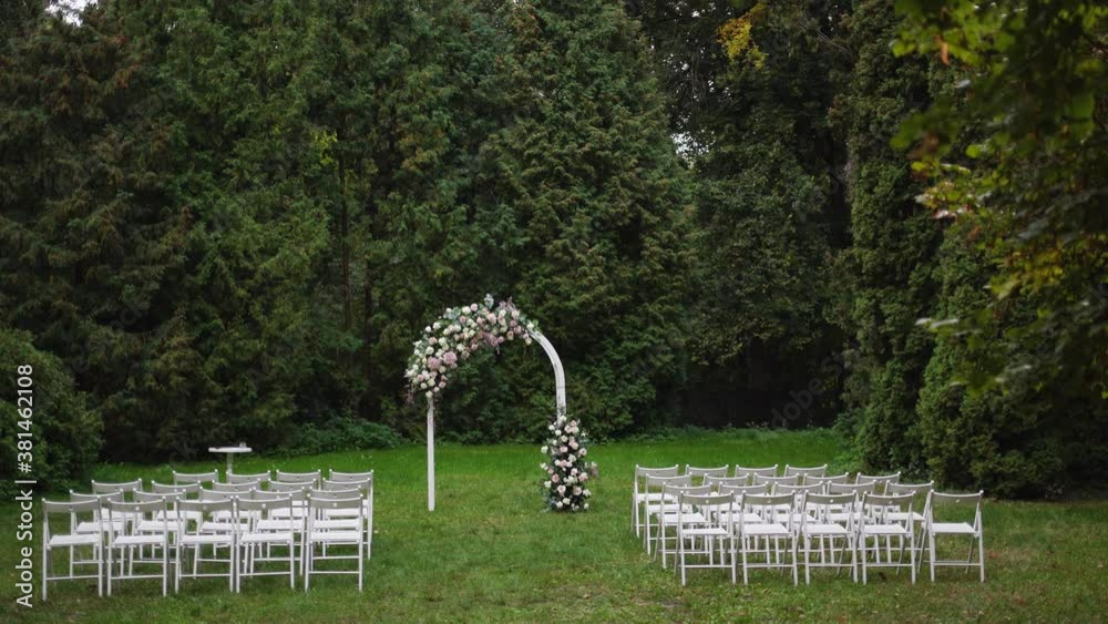 Beautiful wedding floral decorations of flowers in pastel faded colors, wooden painted white frame for outside wedding ceremony in park, raws of many empty chairs standing on green grassy lawn. Beautiful wedding floral decorations of flowers in pastel faded colors, wooden painted white frame for outside wedding ceremony in park, raws of many empty chairs standing on green grassy lawn.
