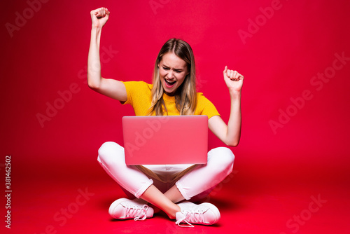 Happy young woman with winning gesture sitting on the floor with crossed legs and using laptop on red background. © dianagrytsku
