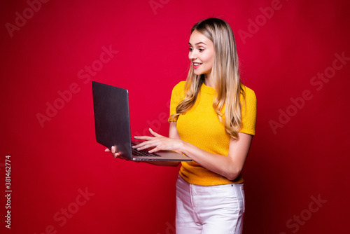 Portrait of smiling young woman holding, working on laptop pc computer isolated on red background.
