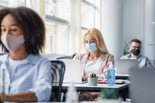 Modern workplace of manager in interior of office. Millennial blonde woman in protective mask works at laptop in office  focus on antiseptic on table