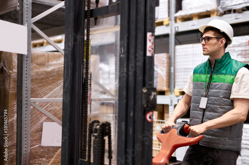 confident warehouse worker with hand pallet truck or pallet jack and the shipment in store, caucasian male in uniform at work