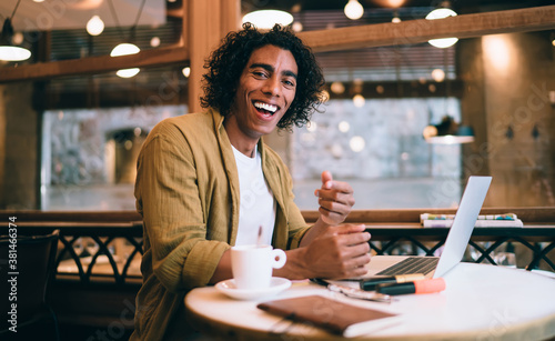 Portrait of prosperous happy curly male freelancer enjoying remote job making research online on laptop computer, young handsome 20s man student enjoying learning courses via technology in cafeteria