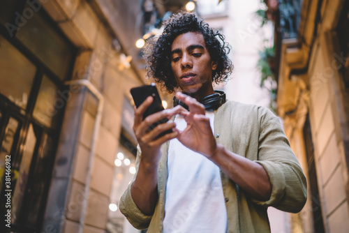 Pensive curly male holding mobile phone sending text messages in social networks chatting online, serious millennial hipster guy holding smartphone making banking via application standing on street