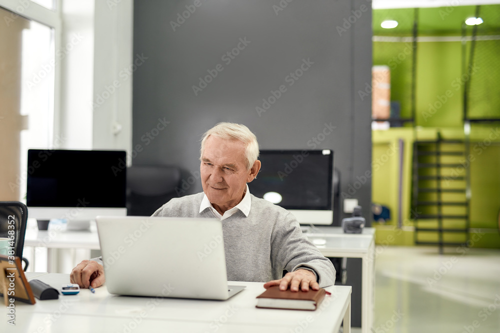 Portrait of cheerful aged man, senior intern looking focused at the screen while using laptop, sitting at desk, working in modern office Portrait of cheerful aged man, senior intern looking focused at the screen while using laptop, sitting at desk, working in modern office