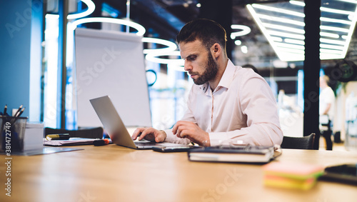 Caucasian male employee reading web information on browsed site survey during productive working process at office table, skilled businessman using 4g internet for making online banking and booking