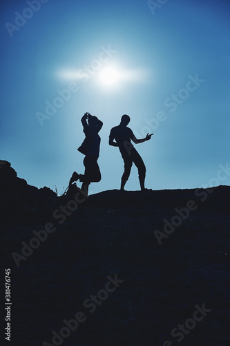 Dark silhouettes of a young couple of a guy and a girl of travelers on the top of the mountain indulge and rejoice against the background of the sun