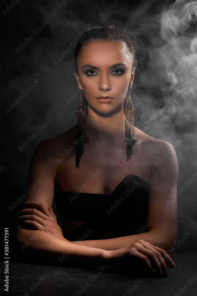 portrait of beautiful sexy brunette woman in fashionable earrings looking at the camera on dark background with smoke