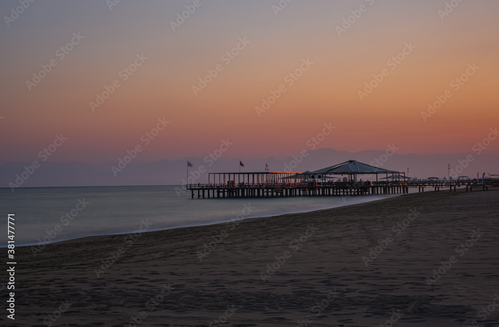 Sunset over the massive concrete pier leading to horizon surrounded by Mediterranean sea. Long exposure shot made in Belek, Turkey. August 2020