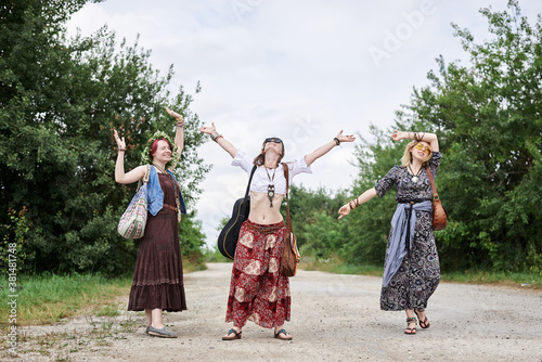 Three hippie women, wearing boho style clothes,, walking on dirt road in countryside, dancing, relaxing, having fun. Friends, traveling together in summer. Freedom and happiness concept.