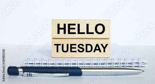 The text Hello Tuesday on a bar of jenga wood, lying on a Notepad with a metal blue pen