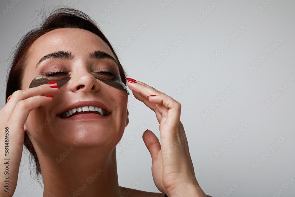 Obraz premium Close-up portrait of attractive young woman applying hydrating patches on her face and smiling. Space for text.
