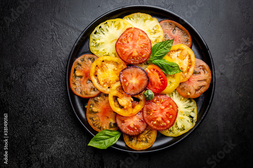 Ripe fresh colorful tomatoes salad with olive oil and basil leaves on black background, top view