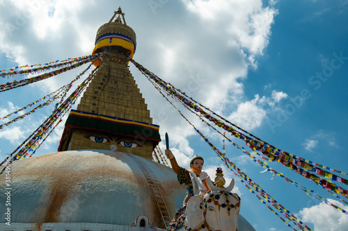 The upper part of the Boudhanath stupa in Kathmandu, Nepal. The image of Buddhas eyes on a gold surface of the stupa. Colorful prayer flags nearby. Buddha statue on a white elephant.