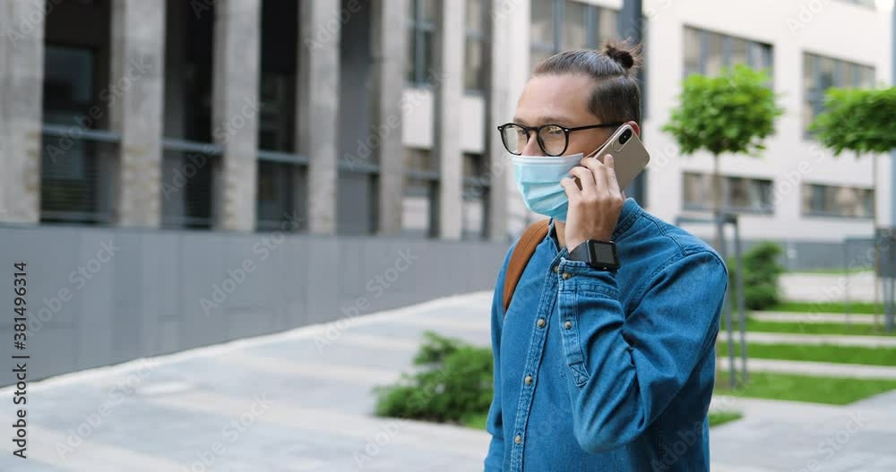 Caucasian young man in glasses and medical mask walking the street and talking on mobile phone. Handsome male in eyeglasses speaking the cellphone outdoor. Telephone call conversation. Caucasian young man in glasses and medical mask walking the street and talking on mobile phone. Handsome male in eyeglasses speaking the cellphone outdoor. Telephone call conversation.
