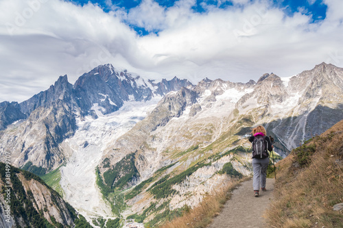 Woman with backpack walking on a trail at high altitude, in Italian alps. High peaks with glaciers and clouded sky in the background