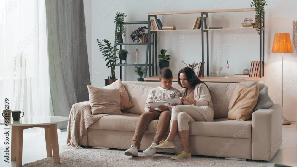 Tracking shot of woman and boy sitting on couch in cozy living room and talking while looking at photos together Tracking shot of woman and boy sitting on couch in cozy living room and talking while looking at photos together