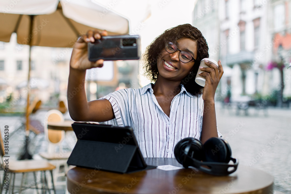 Charming young African dark skinned woman in striped shirt, making selfie photo or having video call, while sitting in outdoor city cafe, drinking coffee and using digital tablet Charming young African dark skinned woman in striped shirt, making selfie photo or having video call, while sitting in outdoor city cafe, drinking coffee and using digital tablet