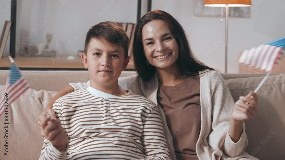 Portrait of happy patriotic woman and boy sitting on couch and waving american flags while looking at camera Portrait of happy patriotic woman and boy sitting on couch and waving american flags while looking at camera