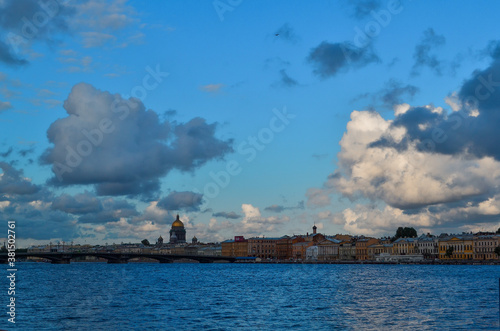 Embankment of Saint Petersburg with colorful historic houses in shade on banks of blue Neva River. Dark and light cumulus clouds in the light of setting sun.