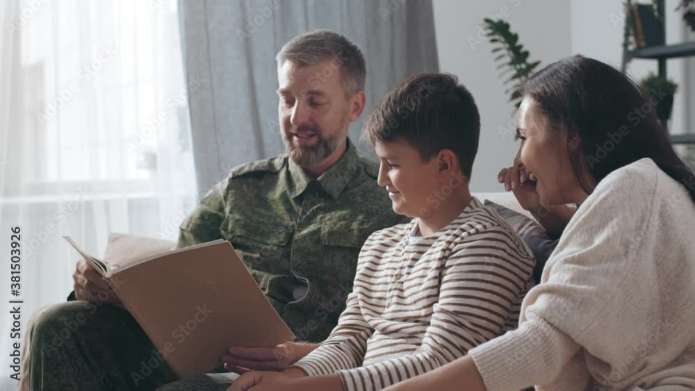 Medium shot of male army officer in military uniform sitting on couch and reading deployment journal with his son and happy wife Medium shot of male army officer in military uniform sitting on couch and reading deployment journal with his son and happy wife