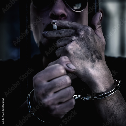 Close up portrait of arrested and handcuffed man imprisoned for crime smokes from behind bars. Gangster concept. © zwiebackesser