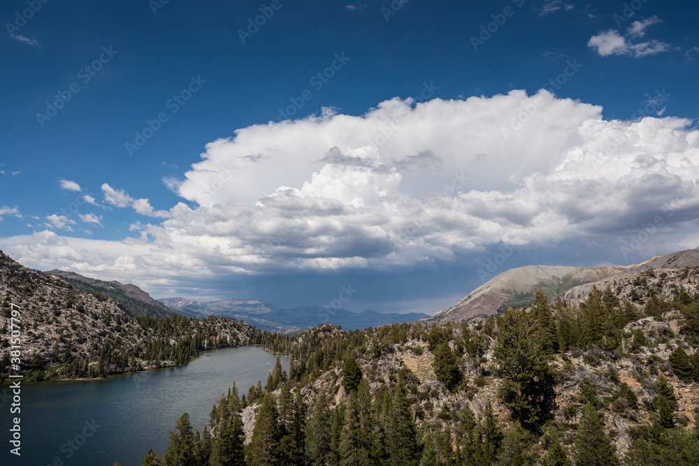 An alpine lake with trees, mountains and dramatic, stormy clouds on a sunny day.