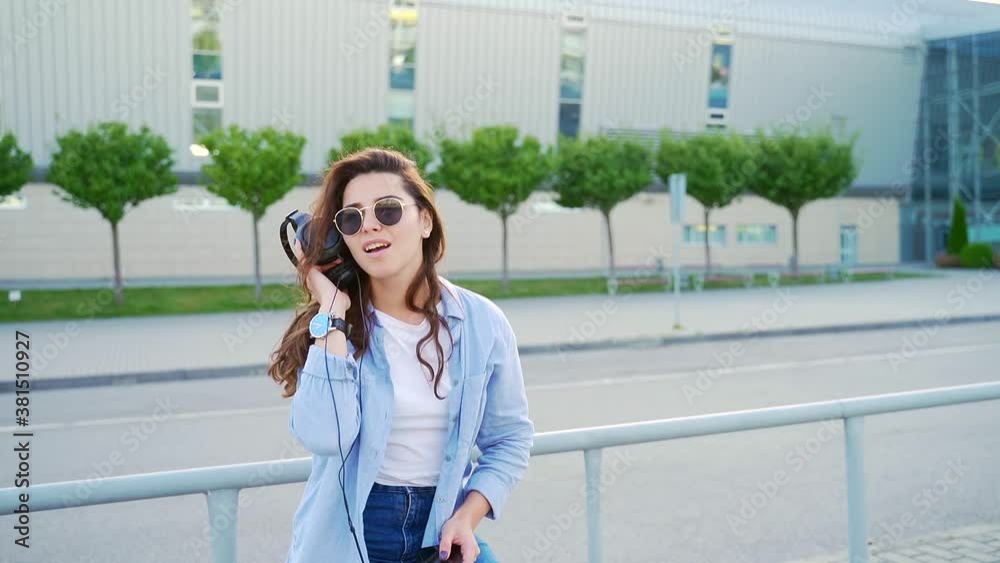 Portrait of cheerful young woman using headphones and modern smartphone for listening music on street. Smiling brunette wearing casual clothing and sunglasses. Portrait of cheerful young woman using headphones and modern smartphone for listening music on street. Smiling brunette wearing casual clothing and sunglasses.