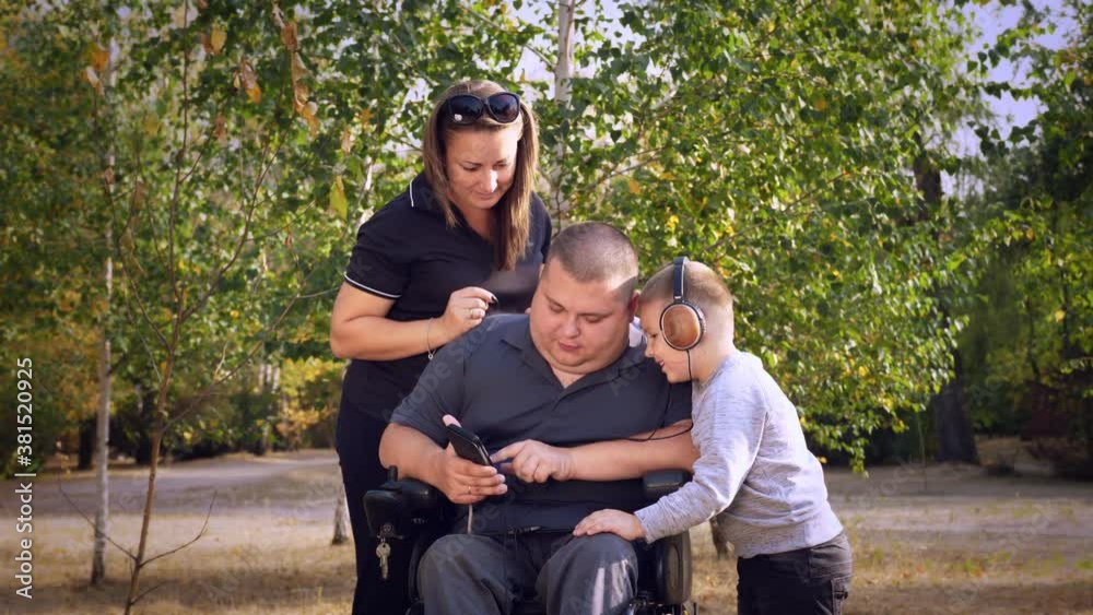 wheelchair man. Handicapped man. young disabled man sits in wheelchair, surrounded by his family, wife and little son. they listen to music, watch videos in smartphone, relax. in city park, on autumn wheelchair man. Handicapped man. young disabled man sits in wheelchair, surrounded by his family, wife and little son. they listen to music, watch videos in smartphone, relax. in city park, on autumn