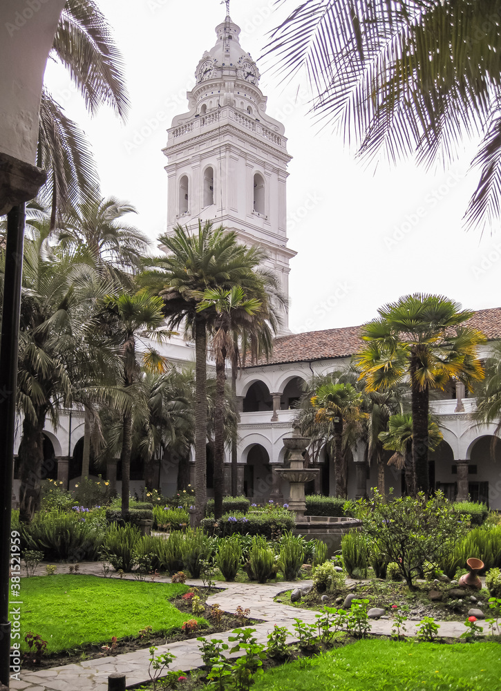 Quito, Ecuador - December 2, 2008: Historic downtown. Inner courtyard with palm trees, scrubs and green garden of Santo Domingo convent. White Church tower. All under silver sky.