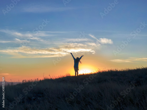 Young inspired Dreadlocks woman raises her hands up standing on the top of a mountain against beautiful sunrise sky. Watching the sunset with beautiful landscape