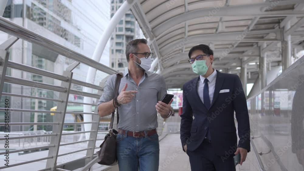 Business people Caucasian with Asian man office worker colleague wearing face mask for protect covid-19 virus walking together on office district and discussing business project with using smartphone. Business people Caucasian with Asian man office worker colleague wearing face mask for protect covid-19 virus walking together on office district and discussing business project with using smartphone.